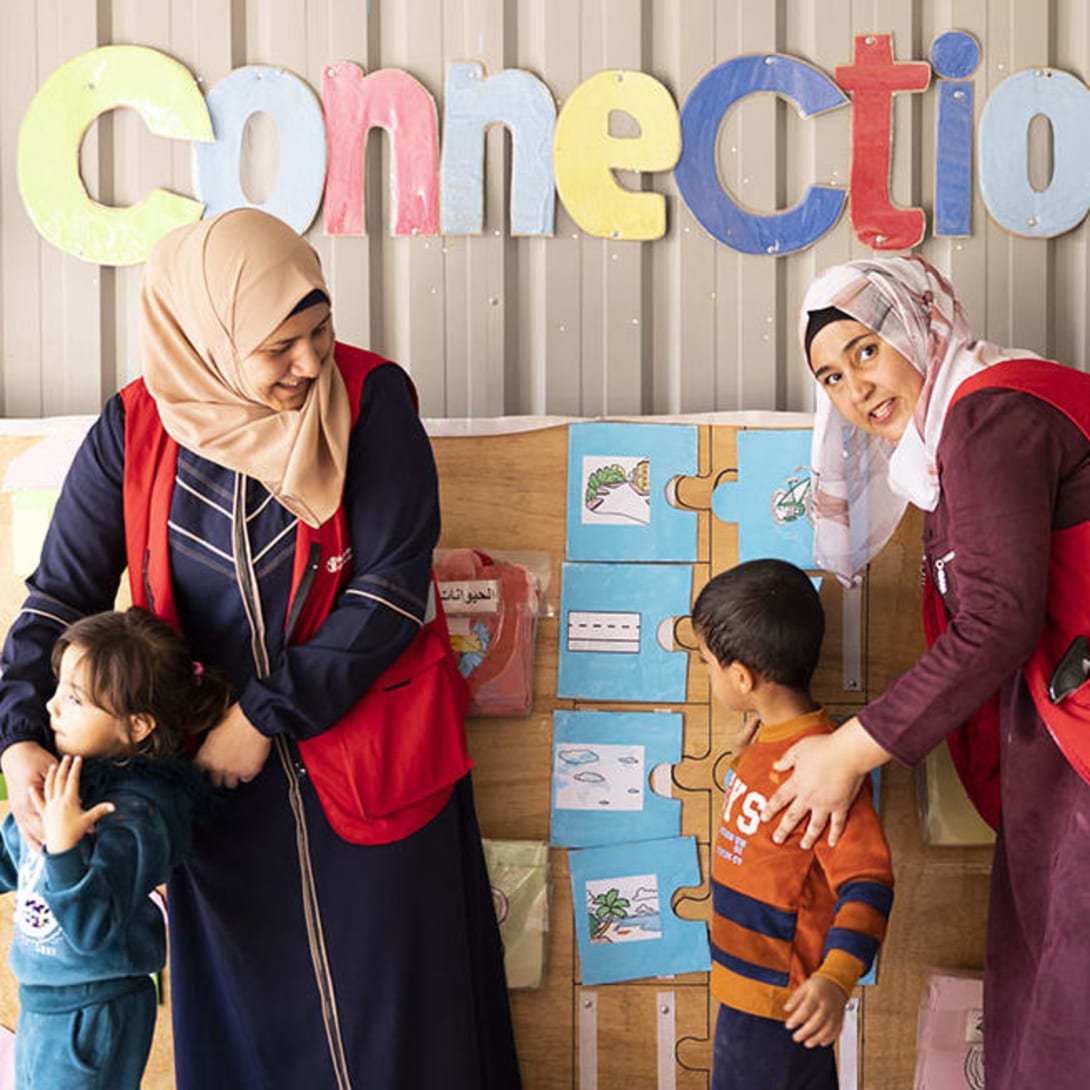 Woman from behind wearing a red Save the Children t-shirt and holding a little girl in her arms.