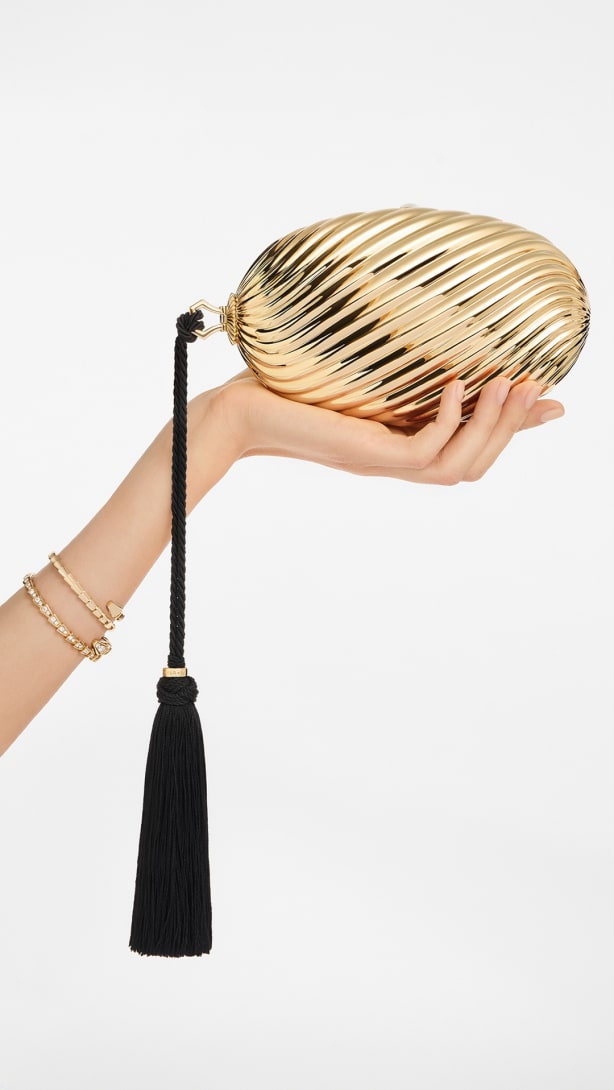 Hand holding the Bvlgari Melone clutch in pleated gold-finish aluminium with black tassel.