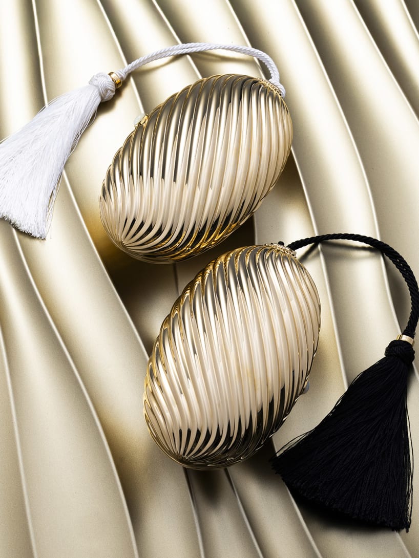 Two gold-finish Melone clutches with black and ivory tassels against a sculptural, wavy backdrop.