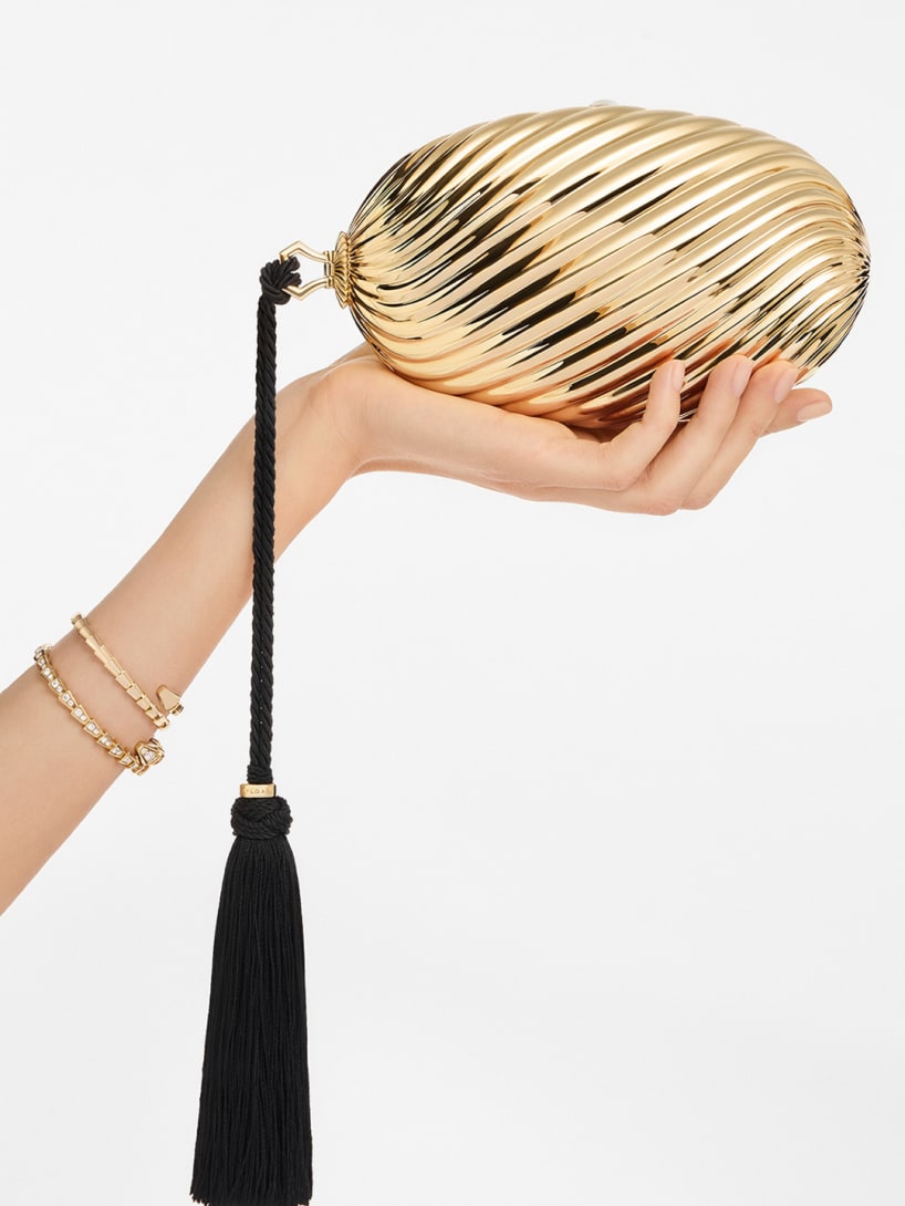 Hand holding the Bvlgari Melone clutch in pleated gold-finish aluminium with black tassel.