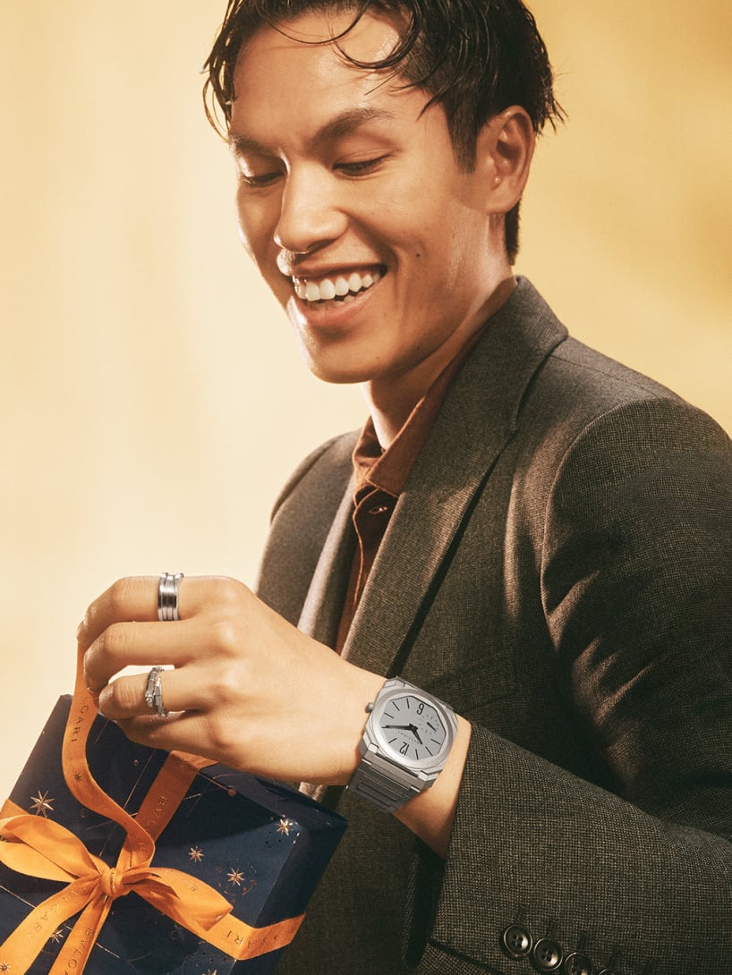 Model wearing Bvlgari jewellery and an Octo Finissimo watch, and holding a gift box, golden backdrop.