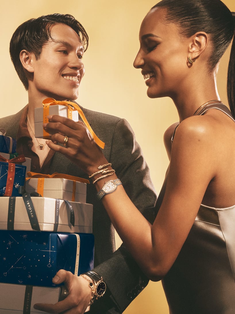 Woman wearing Bvlgari jewellery taking a gift from a man holding Bvlgari boxes, gold backdrop.