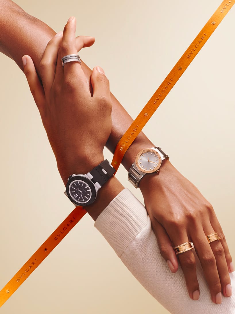 Intertwined hands with orange ribbon, wearing Bvlgari Aluminium and Bvlgari Bvlgari watches, and rings.