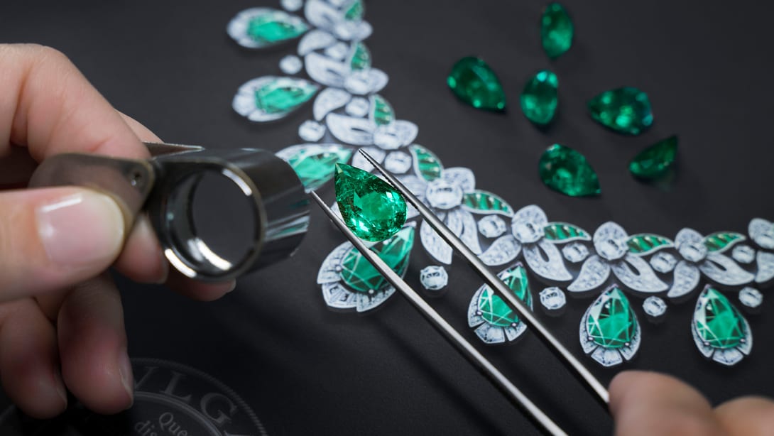 Craftsman looking at an emerald through a lens with sketch of a necklace and emeralds in the background.