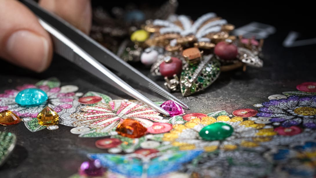 Craftsman placing a coloured gem with a tweezer on the sketch of a Bulgari necklace with flowers, close up.