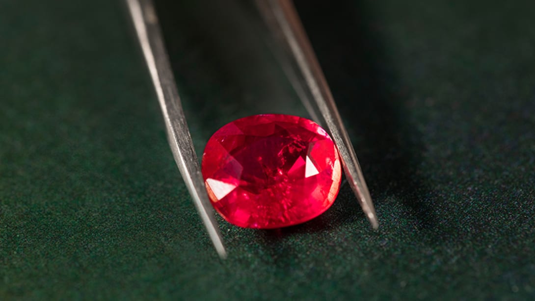 A Bulgari cushion-cut ruby held by tweezers, dark background, close up.