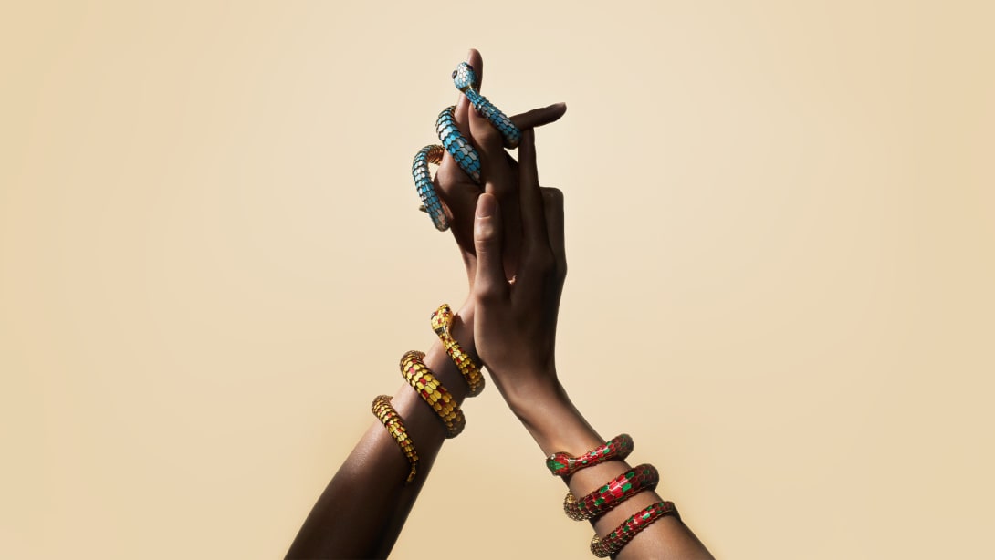 Two Serpenti heritage secret watches with enamel scales coiling around two hands, beige background.